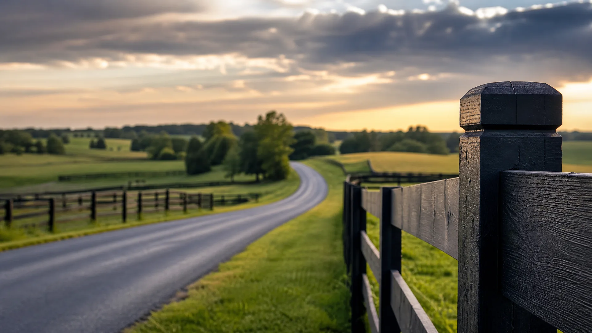 A narrow paved road curves through green pasture at sunset, framed by a dark wooden post-and-rail fence with a close-up corner post on the right.